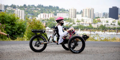 Person in a pink helmet riding a three wheel hand cycle with city skyline in background.